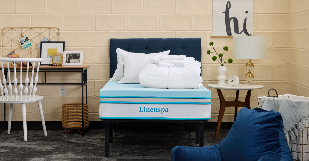A staged bedroom scene with a navy upholstered headboard, a Linenspa mattress topper on a bed frame, white pillows and a folded comforter, and no people.