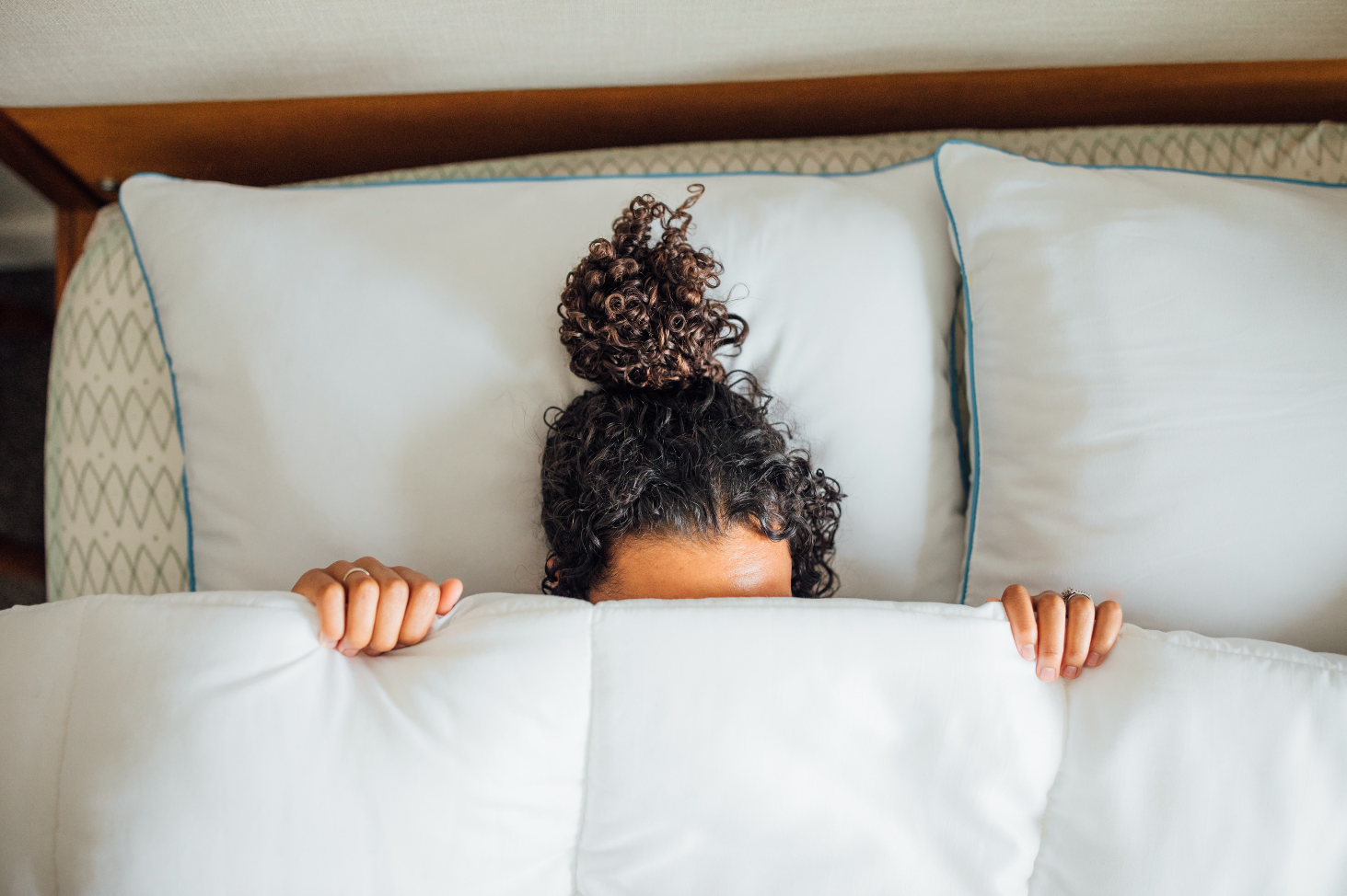 Person with curly hair peeking out from behind pillows on a bed
