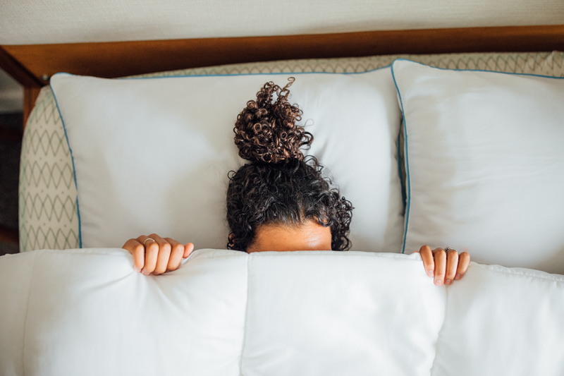 Person with curly hair peeking out from behind pillows on a bed
