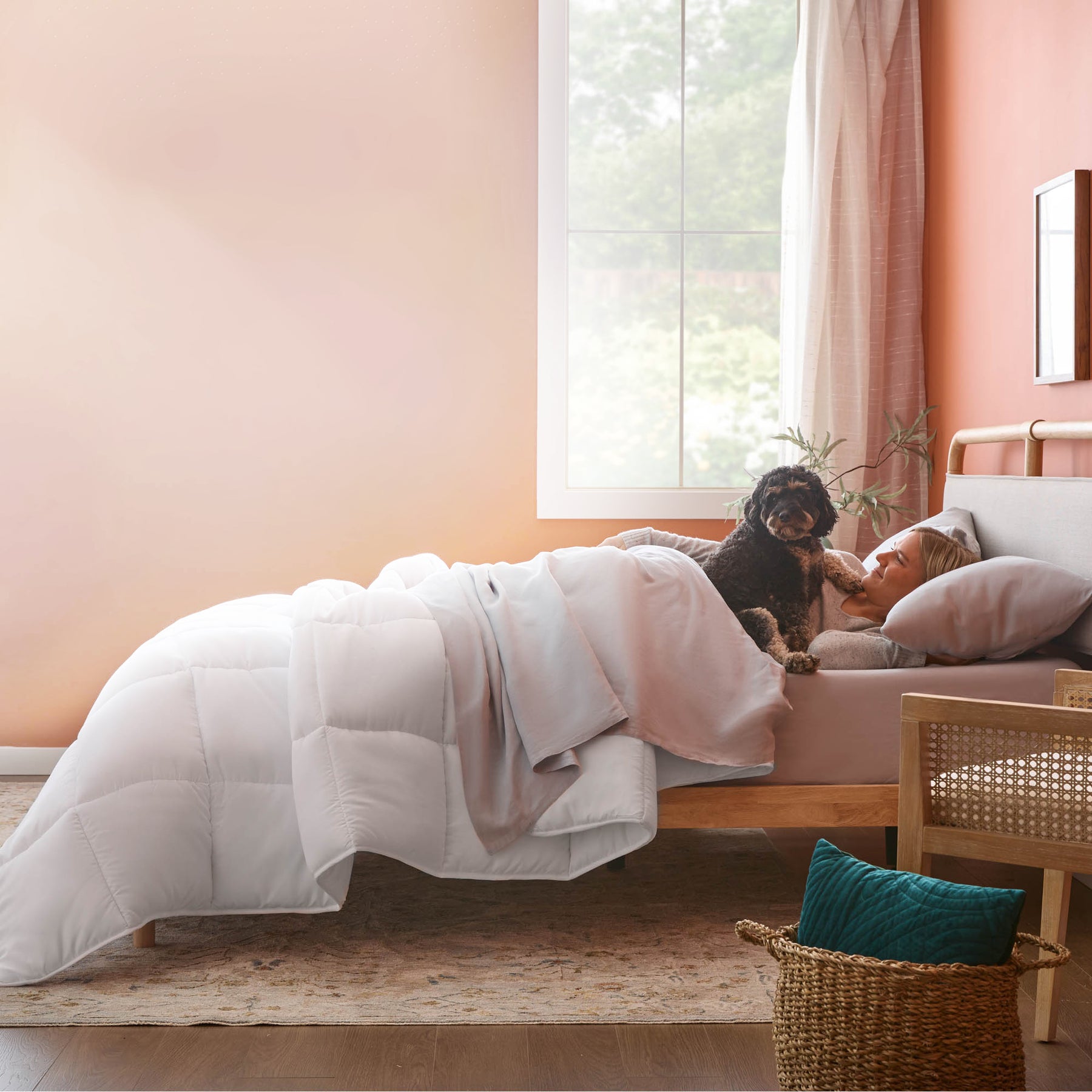 Woman lying on a bed with a dog, surrounded by soft bedding and decor in a cozy bedroom.